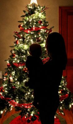 a mother and her child standing in front of a christmas tree with the lights on