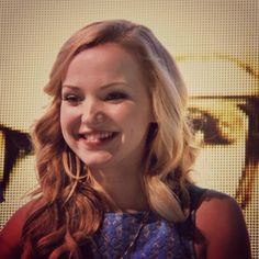 a woman with long blonde hair smiling at the camera while standing in front of a screen