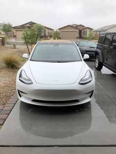 a white tesla parked in a driveway next to two black trucks on the side of the road