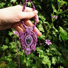 a hand holding a purple beaded necklace in front of green plants and flowers on the other side