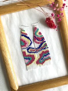 a pair of beaded earrings sitting on top of a wooden frame next to pink flowers