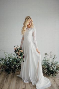 a woman standing in front of flowers wearing a long sleeved wedding dress with sheer sleeves