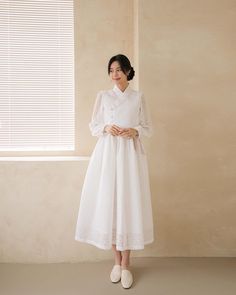 a woman standing in front of a window wearing a white dress with long sleeves and buttons