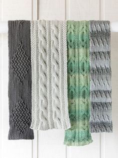 four knitted scarves hanging from a hook on a white wooden wall, all in different shades of green and grey