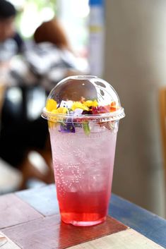 a plastic cup filled with colorful liquid and flowers on top of a wooden table next to a person