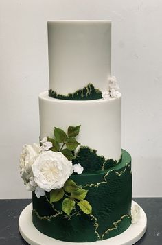 a three tiered cake with white flowers and green leaves on the top, sitting on a plate