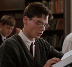 a young man in glasses reading a book while others look at the books behind him