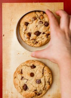 two chocolate chip cookies sitting on top of a cookie sheet next to a hand reaching for one