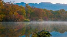 a body of water surrounded by trees in the fall with mist rising from it's surface