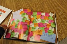 an open book with sticky notes attached to it on a wooden table next to a person's hand