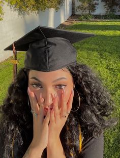 a woman wearing a graduation cap covers her face with her hands while sitting in the grass