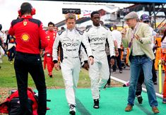 two men in racing suits walk down the green carpet as people watch from behind them