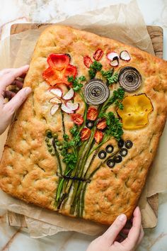 a square pizza with vegetables and olives on it is being held by two hands