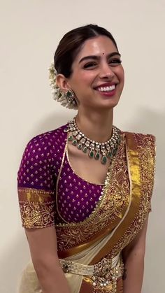 a woman in a purple and gold saree smiles at the camera with her hand on her hip