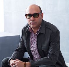 a man sitting on top of a white chair wearing sunglasses and a black jacket with his arms crossed