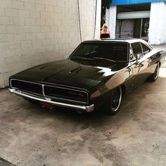 a black muscle car parked in front of a garage with a woman standing next to it