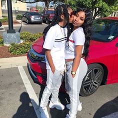 two girls standing next to each other in front of a red car on the street