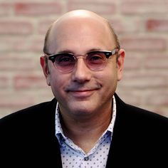 a bald man wearing glasses and a polka dot shirt smiles at the camera while standing in front of a brick wall