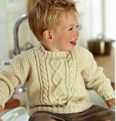 a young boy sitting on top of a kitchen counter wearing a sweater and pants, smiling at the camera