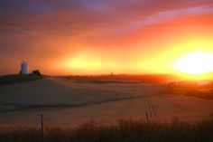 the sun is setting over a field with a light house in the distance and clouds