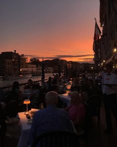 people are sitting at tables in front of the water as the sun is setting over some buildings