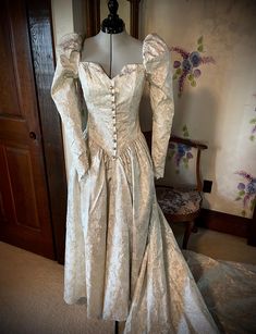 This renaissance-style dress is truly unique. The fitted princess line bodice has a square neckline front and back with a small vee at center front. This is accentuated by a mock opening in the front with "pearl" buttons, each encircled with rhinestones. Some of the buttons are missing a rhinestone. Puff sleeves, with elastic at the shoulders for wearing ease, taper to points at the wrist with a four button and loop closure. The drop waist come to a point in the front and back. Skirt is gathered. Zipper closure. What makes this truly renaissance looking is the fabric. It has gold metallic slightly raised flowers on a cream background. Dress has been cleaned and preserved since the 1991s and is in excellent condition.  Not only would this make a wonderful wedding dress, it would also be ama Elegant Gown With Fitted Bodice And Heart-shaped Neckline, Elegant Ball Gown With Lined And Fitted Bodice, Formal Gown With Fitted Bodice And Sweetheart Neckline, Formal Gown With Sweetheart Neckline And Fitted Bodice, Ball Gown With Boned And Fitted Bodice, Boned Bodice Fitted Ball Gown, Fitted Ball Gown With Ruched Bodice And Sweetheart Neckline, Fitted Gown With Heart-shaped Neckline For Debutante Ball, Heart-shaped Neckline Gown With Fitted Bodice For Debutante Ball