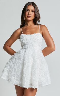 a woman wearing a white dress with flowers on the skirt and spaghetti top, standing in front of a gray background