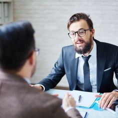 a man sitting at a table talking to another man who is wearing glasses and a suit