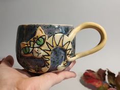 a hand holding a ceramic mug with a cat on it's side and leaves in the background