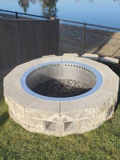 an outdoor fire pit sitting on top of grass next to a fence and water in the background