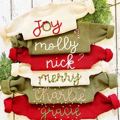 christmas sweaters are hung on a white wooden wall with green garland and pine cones