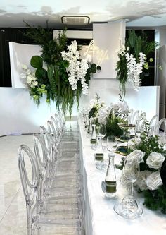 a long table with white flowers and greenery in vases on the top is surrounded by clear acrylic chairs