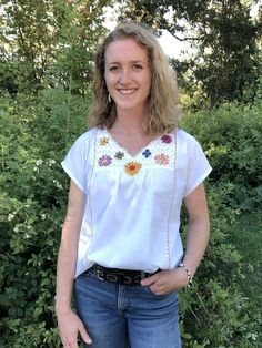 a woman standing in front of some bushes wearing jeans and a white shirt with flowers on it