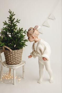 a baby in white is looking at a small christmas tree that has been placed on a chair