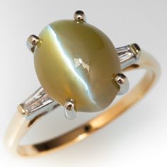 This wonderful 14K yellow gold ring is centered with one (1) oval cabochon cut natural cat's eye chrysoberyl weighing 6.29 carats and set into am 18K white gold four-prong head. The shoulders of the ring are each accented with one (1), channel set, tapered baguette cut diamond. The ring measures 10.8mm at the top, rises 11.3mm above the finger, tapering to 1.6mm wide and 1.1mm thick at the base of the shank. This ring is currently a size 7.25. Antique Cocktail Ring, Cats Eye Ring, Natural Cat, Baguette Cut Diamond, Channel Set, Baguette Cut, Yellow Gold Ring, Oval Cabochon, Yellow Gold Rings