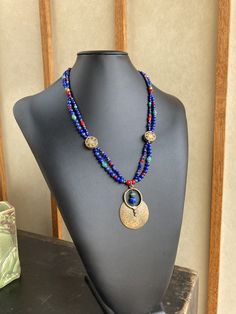 Featuring stones that are associated with wisdom, insight, spirituality, and communication, this is made with gorgeous deep royal blue to navy blue grade A Lapis in various shapes and sizes, which contrast strikingly with green semi precious turquoise, rich red Czech glass and white-heart beads, and brass spacers. The two strands gather at Tibetan brass 15mm coin beads inlaid with red coral and turquoise howlite as well as other brass beads. A 53x37mm plated antique gold large tribal style medal Handmade Bohemian Sapphire Necklace, Bohemian Lapis Lazuli Beaded Necklaces As Gift, Bohemian Lapis Lazuli Beaded Necklaces For Gift, Bohemian Lapis Lazuli Beaded Necklace For Gift, Blue Lapis Lazuli Jewelry For Festivals, Handmade Blue Lapis Lazuli Necklace, Handmade Blue Medallion Necklace, Colorful Beads Lapis Lazuli Necklace Gift, Unique Blue Gemstone Beaded Necklaces