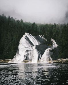 a large waterfall in the middle of a lake surrounded by trees and foggy skies