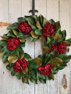 a wreath with red flowers and green leaves hanging on a white wooden door, decorated with an iron hook