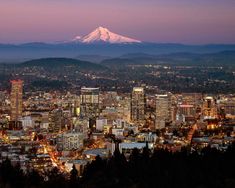 the city lights are on as the snow capped mountain looms in the distance