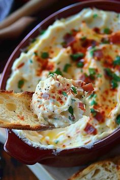 a red bowl filled with mashed potatoes topped with bacon and green chives next to slices of bread