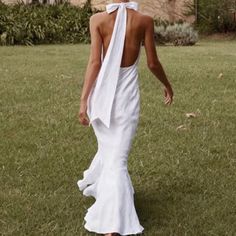 a woman in a white dress walking across a grass covered field with her back to the camera