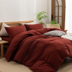 a bed with red comforter and pillows in a room next to a plant on the floor