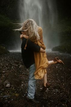 a man and woman hug in front of a waterfall