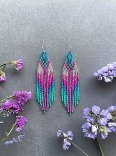 two pairs of beaded earrings sitting next to purple flowers on a gray surface with pink and blue beads