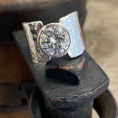 Moissanite Ring Handmade By Wild Prairie Silver Jewelry 3 carat Hearts & Arrows Moissanite set into a asymetrical sterling band. Moissanite is an ethical diamond substitute. Handmade with love in rural Wisconsin by Wild Prairie Silver Follow Us: Instagram: https://www.instagram.com/wildprairiesilver/ Facebook: https://www.facebook.com/wildprairiesilver/ Visit our Jewelry Website www.wildprairiesilver.com Handmade Jewelry Supplies www.savannahjewelrysupply.com Contact Us: Call or Text: 920-36 Formal Jewelry With Tension Setting In White Topaz, Dazzling Tension Set Anniversary Jewelry, Unique Diamond White Jewelry For Anniversary, Unique Jewelry With Tension Setting For Anniversary, Unique Brilliant Cut Diamond Ring Gift, Silver Moissanite Bezel Set Jewelry, Silver Moissanite Jewelry With Bezel Setting, Sterling Silver Diamond Ring With Wide Band As Gift, Modern Moissanite Jewelry With Center Stone