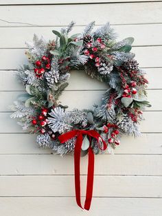 a christmas wreath hanging on the side of a white building with red ribbon and evergreen leaves