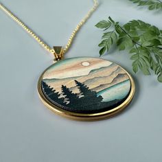 a necklace with an image of mountains and trees on it next to a leafy plant