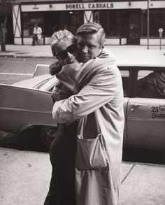 an old man and woman hugging on the street in front of a car, black and white photograph