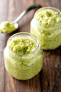 two small jars filled with green pesto on top of a wooden table next to a spoon