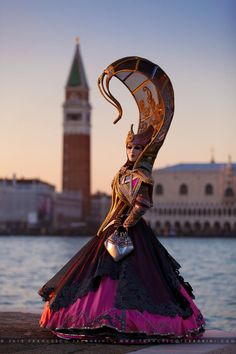 a woman in a long dress is standing near the water with a large clock tower in the background